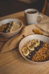 Warm morning setup with two bowls of oatmeal topped with banana, chia, dried berries, and granola, served with tea and golden spoon on wooden table with waffle cloth