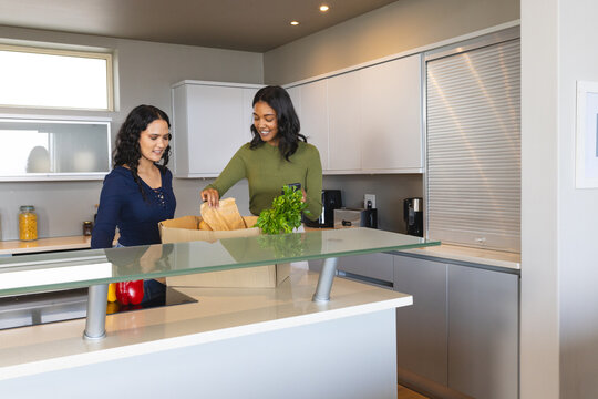 Diverse female friends unpacking groceries at kitchen island, with grocery bag, parsley, smartphone