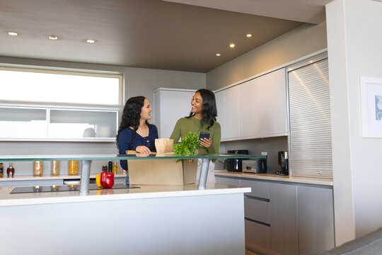 Smiling Diverse female friends unpacking groceries at kitchen island, with bell pepper and lettuce