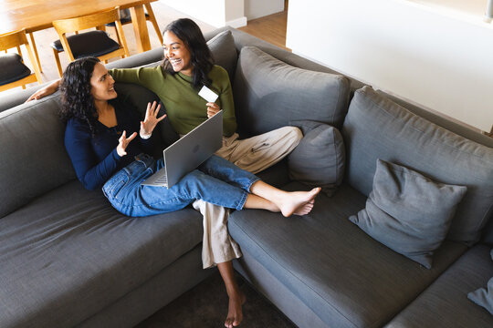 Sitting Diverse female friends browsing online store on laptop in living room, holding credit card