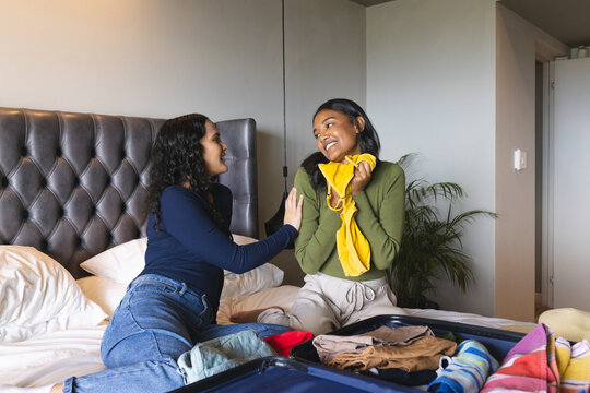 Laughing diverse female friends sorting clothes on bed in bedroom, with open suitcase - Powered by Adobe