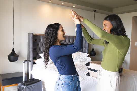 High-fiving diverse female friends sharing joyful gesture in modern hotel room, with suitcases