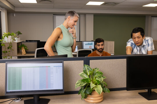 Diverse coworkers examining spreadsheets on monitors in open-plan office, with plants