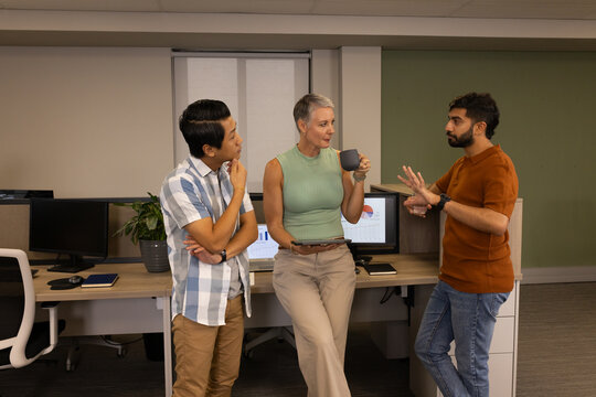 Diverse coworkers discussing quarterly reports in open office, with tablet coffee mug and monitors