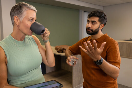Chatting Diverse coworkers standing at office break counter, with tablet and dark coffee mug