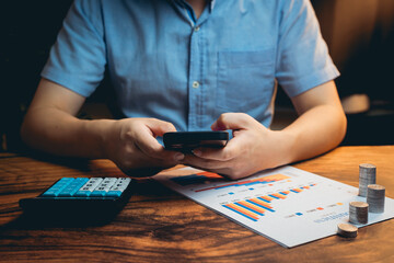 Person Analyzing Financial Data with Smartphone and Calculator on Wooden Desk Amidst Graphs and...
