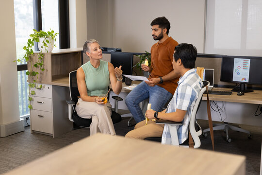 Holding bananas and apple, Diverse coworkers collaborating in open-plan office, with monitors
