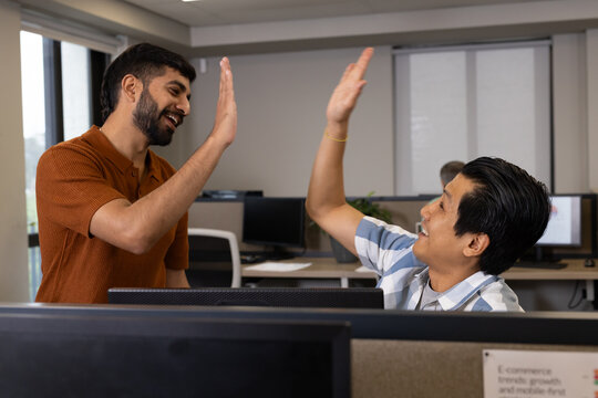 High-fiving Diverse male coworkers celebrating success in open-plan office, with computer monitors