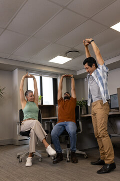 Stretching Diverse coworkers raising arms overhead in office space, with rolling chairs and desks