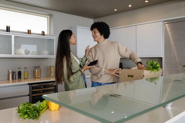 Smiling Diverse couple unpacking groceries from box at kitchen island, with smartphone and herbs