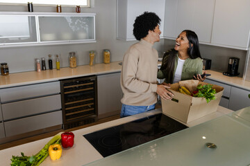 Laughing Diverse couple unpacking cardboard grocery box in modern home kitchen, with smartphone