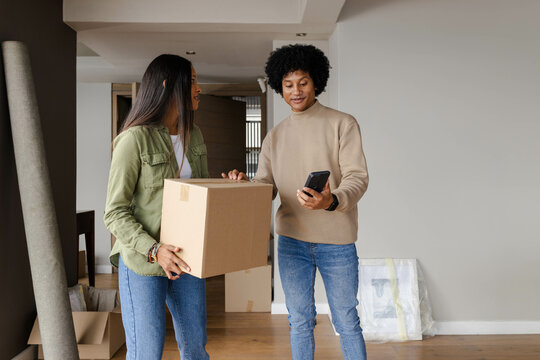 Diverse couple holding moving box and checking smartphone in living room, with rolled-up carpet