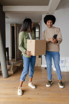 Unpacking Diverse couple organizing moving boxes in empty room, with smartphone and rolled-up rug