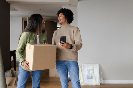 Laughing Diverse couple holding moving box and smartphone in empty living room, with bubble wrap