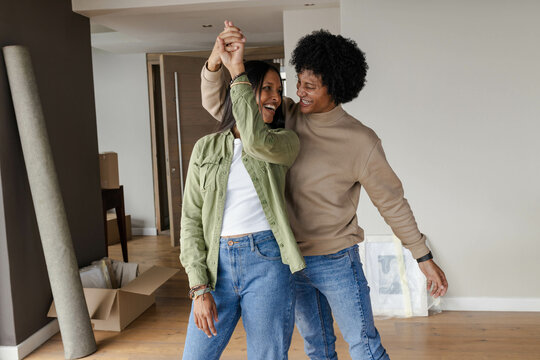 Smiling diverse couple dancing in new home, with cardboard box and wrapped picture frames