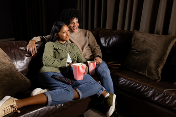Snuggling diverse couple on leather sofa in home theater, holding striped popcorn tubs