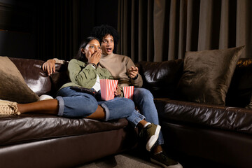 Watching diverse couple reacting with surprise on leather couch at home, holding popcorn buckets