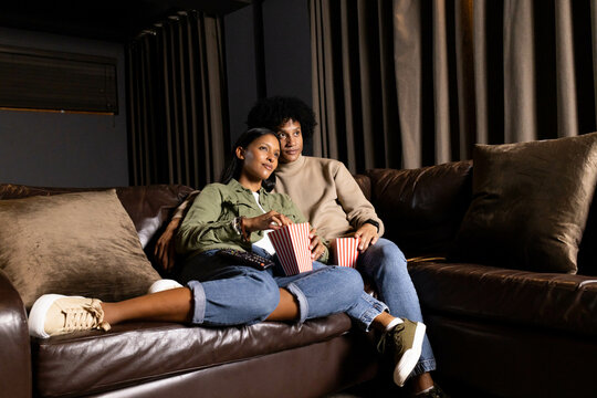 Diverse couple sitting on leather couch in dimly lit home theater, holding striped popcorn buckets - Powered by Adobe