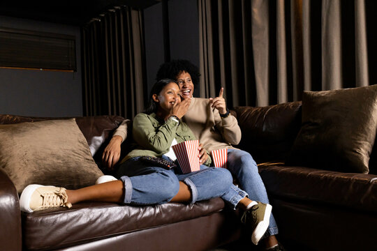 Reacting Diverse couple covering mouth and pointing on sofa at home, with striped popcorn buckets - Powered by Adobe