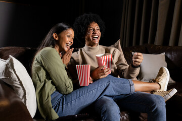 Laughing diverse couple holding red-and-white striped popcorn buckets on brown couch in living room