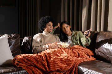 Cuddling diverse couple holding popcorn and remote on leather couch in living room, with blanket