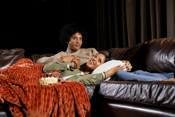 Lounging diverse couple relaxing on leather sofa in dimly lit living room, with popcorn bowl