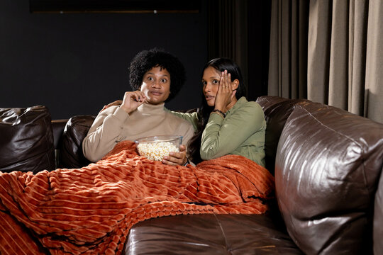 Reacting Diverse female friends holding popcorn bowl on couch in dim living room, with orange throw