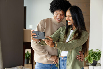Smiling Diverse couple taking selfie beside wooden panel in modern living room, with smartphone