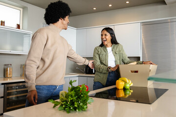Smiling Diverse couple unpacking cardboard grocery box at kitchen island, featuring fresh produce