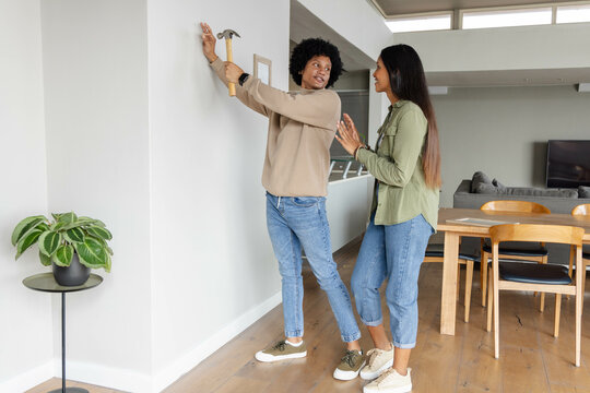Diverse couple hanging picture frame on wall in open-plan living area, with hammer and plant