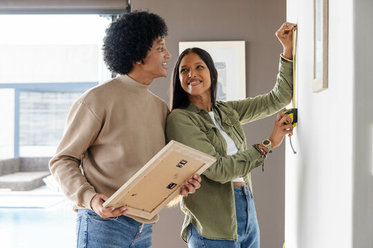 Using yellow tape measure, diverse couple hanging framed picture on neutral wall in living room - Powered by Adobe