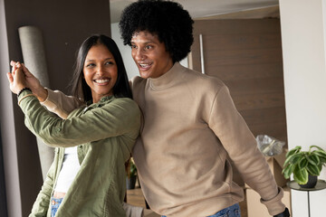 Dancing diverse couple guiding each other's arms in living room, with moving boxes and potted plant