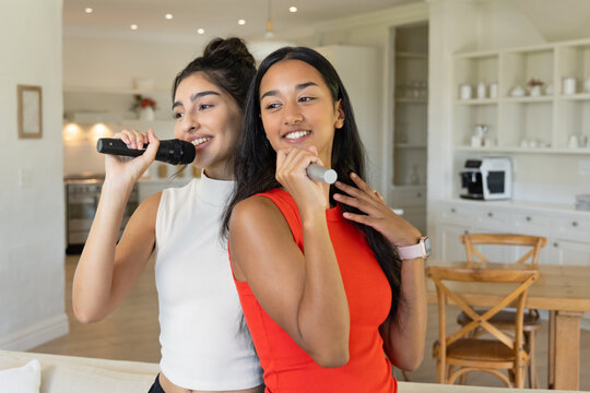 Standing Diverse female friends singing karaoke in modern kitchen dining area, with microphones