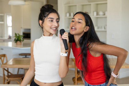Singing diverse female friends standing in modern home kitchen and dining area, with microphone