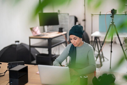 A woman wearing a hijab is focused on her work, typing on a laptop and writing notes. She is in a studio environment, possibly editing or planning creative content.