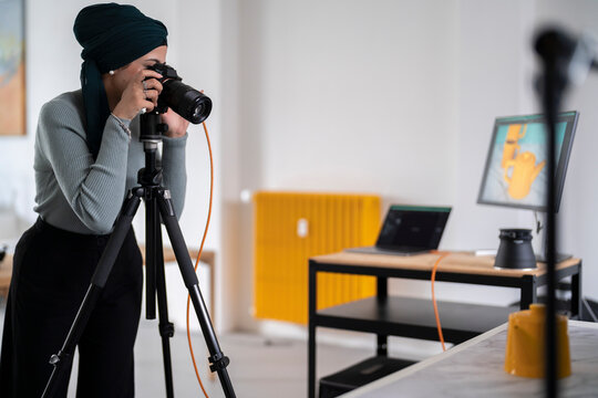 A woman wearing a headscarf is using a camera on a tripod to take photos in a studio setting. She is engaged in professional photography work, likely for a project.