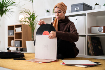 A woman wearing a hijab carefully removes a vinyl record from its sleeve. She is getting ready to enjoy listening to music on her turntable, appreciating the warm sound of analog audio at home.