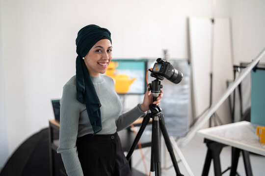 A woman in a headscarf stands in a studio, holding a camera on a tripod. She is a photographer setting up for a shoot, ready to capture images in her workspace.