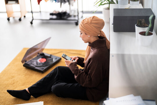 A woman in a headscarf sits on the floor, scrolling on her phone while a record spins on a nearby player. She is likely relaxing and enjoying some music and screen time at home.