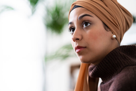 A woman wearing a brown hijab and sweater looks thoughtfully into the distance. She appears to be contemplating something important or observing her surroundings with focus.
