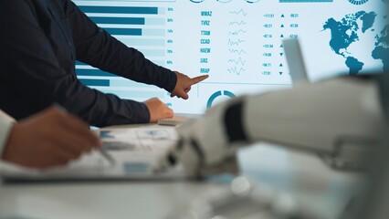 Professionals engage in data analysis, utilizing a robotic hand and digital screen to navigate financial trends and insights during a collaborative workplace discussion.Noogenesis