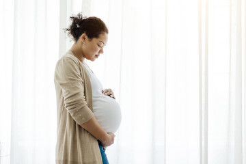 Profile Portrait Of Smiling Black Pregnant Woman Tenderly Embracing Belly Against Window At Home, Happy Expectant Mother Enjoying Her Healthy Pregnancy And Awaiting Time, Side View With Copy Space © Anastasiya