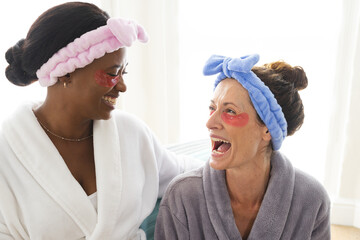 Laughing Diverse female friends sitting on sofa in home spa lounge, with red under-eye gel patches
