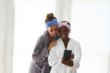Diverse female friends checking smartphone in spa, wearing robes, headbands and under-eye patches
