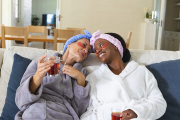 Relaxing diverse senior female friends holding tea mugs on sofa at home, with under-eye gel patches