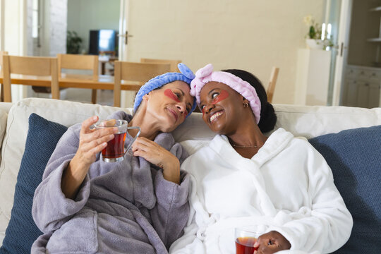 Relaxing diverse senior female friends holding tea mugs on sofa at home, with under-eye gel patches - Powered by Adobe