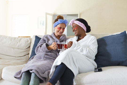 Sipping tea Diverse female friends wearing bathrobes and under-eye patches on living room sofa - Powered by Adobe