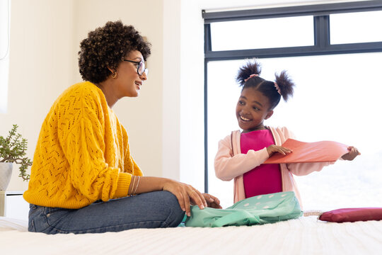Smiling diverse mother and daughter sharing homework on bed in bright bedroom, with pink folder