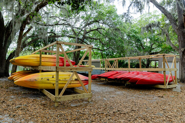 Stacked colorful kayaks on a wooden rack under a large tree near a calm river, outdoor recreation...