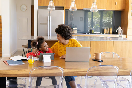 Smiling mother and African American child sitting at dining table in kitchen, with laptop, tablet